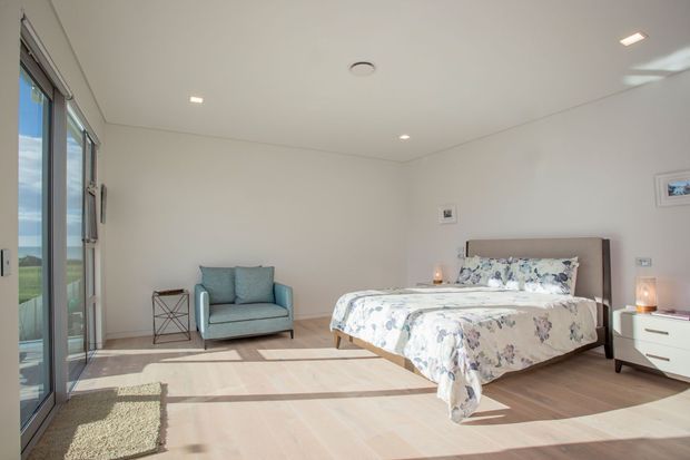 Bright contemporary bedroom with a low-profile upholstered bed, floral bedding, pale timber flooring, white walls, a blue armchair, minimalist bedside furniture, and extensive sliding glass doors opening to a scenic exterior view.