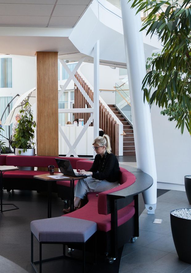 A bright, contemporary multi-level commercial interior with an open stairwell and sculptural white structural supports. The space features a curved burgundy upholstered banquette with integrated dark edging, loose ottomans, small work tables, and a person working on a laptop. A timber-clad structural column, white geometric bracing, glass balustrades, dark tiled flooring, indoor planting, and abundant glazing create a modern collaborative lounge or reception area.