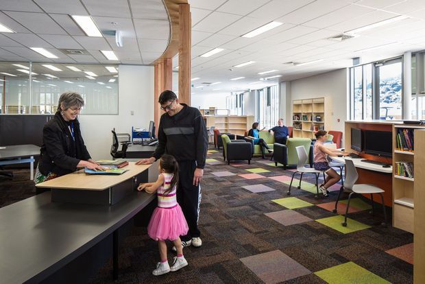 A bright, contemporary library or learning commons with open-plan study areas, computer workstations, built-in timber shelving, lounge seating, and a curved black reception or circulation desk. Several people are using the space, which features patterned carpet tiles, extensive glazing, white acoustic ceiling tiles, and warm timber detailing.