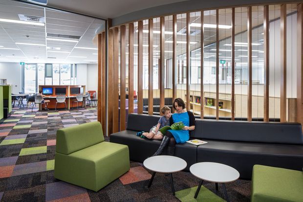 A bright modern library or educational interior with a casual lounge seating area in the foreground. A black upholstered built-in sofa, green armchairs, round white coffee tables, and colourful patterned carpet create an inviting reading and waiting zone. Timber vertical slats divide the lounge from an open study or computer area, with desks, shelving, and extensive ceiling lighting visible beyond.