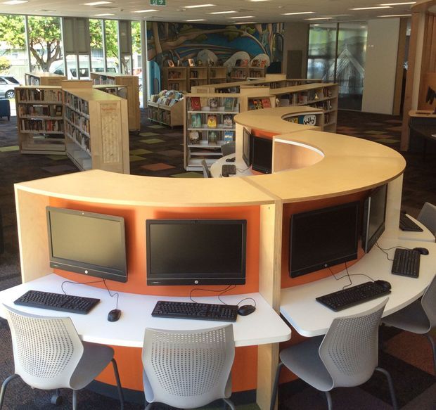 Interior view of a modern library or educational resource centre, featuring a large curved computer workstation with multiple desktop computers, keyboards and ergonomic chairs in the foreground. Low timber bookcases, bookshelves, reading areas, patterned carpet, large windows, and a colourful mural define the space.