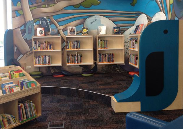 A colourful children's library or reading area with low wooden bookcases filled with books, whimsical blue sculptural reading furniture, floor cushions, patterned carpet, and a large illustrated woodland mural covering the back wall. The space is playful and purpose-built for young children.