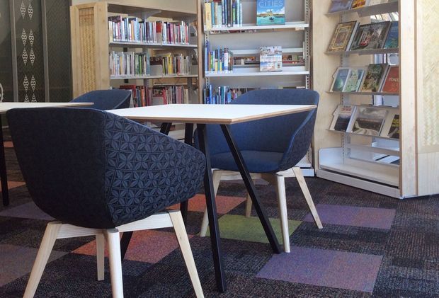 A compact, stylish reading and meeting area with a light timber table, upholstered blue armchairs, full-height bookshelves, and a freestanding magazine display. The space has patterned carpet tiles in muted multicolour tones and decorative patterned wall panels, creating a warm contemporary library or lounge setting.