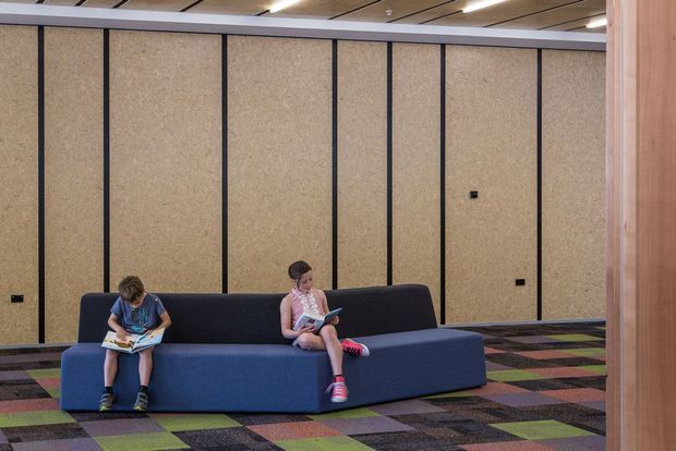 A contemporary communal lounge or waiting area with two children seated on a large modular blue sectional sofa. The space features full-height tan acoustic wall panels divided by dark vertical trims, a multicoloured patterned carpet, and a linear timber-slatted ceiling with recessed lighting. Timber framing or joinery is visible at the right edge.