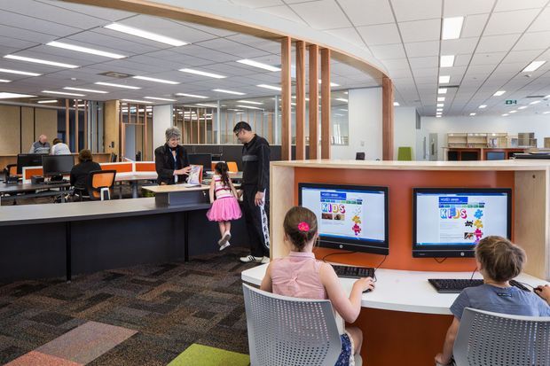 Bright open-plan commercial interior, likely a public library or community learning space, with computer workstations, children using desktop computers, long service counters, bookshelves, and curved timber-framed partitions. The space has a suspended acoustic tile ceiling, patterned carpet, white work surfaces, and warm wood detailing.