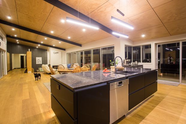 A spacious open-plan kitchen and living area featuring a large dark island with a polished stone benchtop, integrated stainless-steel dishwasher, and contemporary black cabinetry. Warm timber flooring and a timber-panelled ceiling contrast with white walls, exposed dark ceiling beams, pendant and recessed lighting, and extensive glazing. A dining and lounge area is visible beyond the island.
