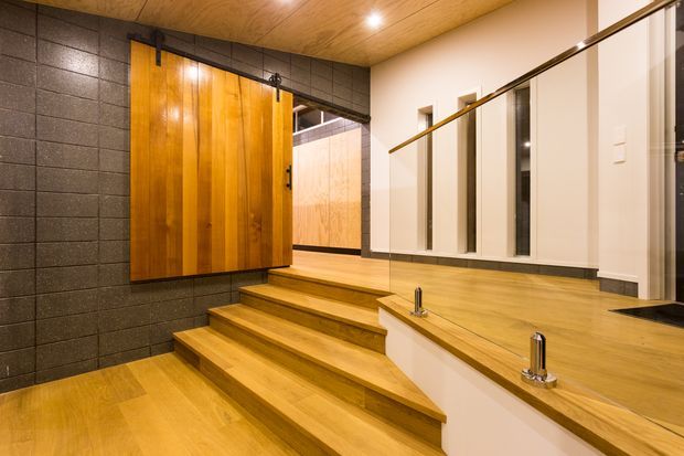 Warm contemporary interior showing a timber-finished stairway beside a long timber vanity with integrated basins and a large mirror. Dark textured masonry tiles contrast with the honey-toned timber ceiling, steps, cabinetry and benchtop, while white walls and recessed lighting brighten the space.