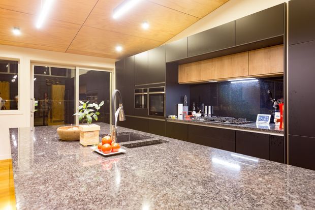 A contemporary kitchen with extensive dark built-in cabinetry, integrated ovens, a gas cooktop and glossy dark splashback. A large polished stone island with an undermount sink dominates the foreground, while warm timber ceiling panels and timber-toned cabinet fronts add contrast. Glazed doors open toward an outdoor area.