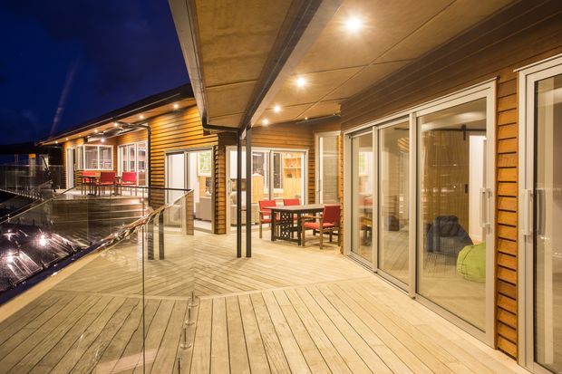 Night-time view of a spacious timber-clad outdoor deck or terrace with glass balustrades, built-in ceiling downlights, and extensive glazed sliding doors opening into warmly lit interior spaces. Outdoor dining furniture is arranged along the deck, which appears to wrap around the building.