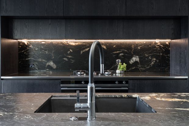 Contemporary dark-toned kitchen viewed from the island, featuring black wood-grain cabinetry, a dramatic dark veined stone splashback and island benchtop, integrated cooking appliances, and a large undermount sink with a tall chrome gooseneck tap. Warm linear under-cabinet lighting highlights the stone surface.