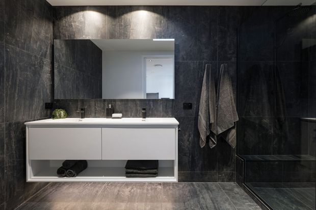 A dark, contemporary bathroom with charcoal stone-look wall and floor tiles, a floating white double vanity with integrated basin and open lower shelving, black tapware and hardware, a large rectangular mirror, and a frameless glass shower enclosure. Warm recessed lighting highlights the textured surfaces.
