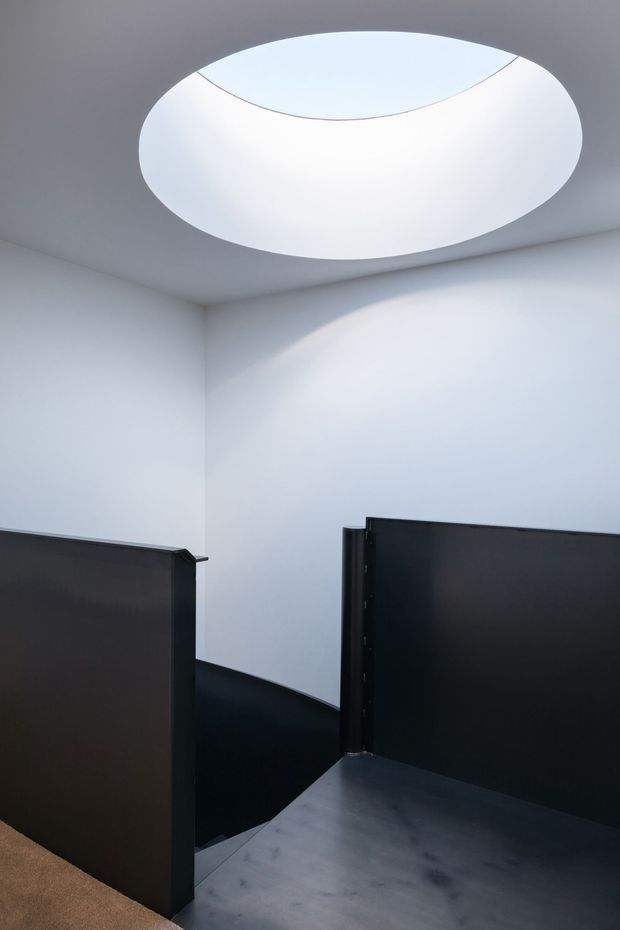 Minimalist interior view of a stairway or landing with dark matte partition panels and a curved stair enclosure. White walls and ceiling are dramatically illuminated by a large circular skylight, creating a bright, sculptural focal point. The lower floor surface appears dark grey and smooth, with a small area of beige carpet visible at the edge.