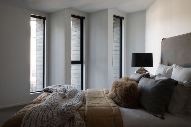 A cozy contemporary bedroom with a large upholstered bed, layered neutral bedding, textured knitted throws, dark cushions, and a black bedside lamp. Three tall, narrow windows with dark frames bring in natural light, while grey walls and soft neutral furnishings create a calm, refined atmosphere.