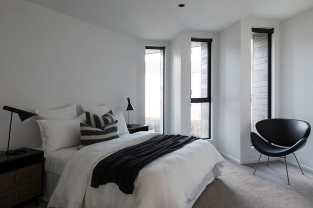 A bright, minimalist bedroom with a white upholstered bed dressed in white bedding and a black throw, dark striped accent cushions, black bedside furniture, and a sculptural black lounge chair. Tall, narrow black-framed windows bring in natural light, while light grey carpet and white walls create a calm, monochromatic interior.