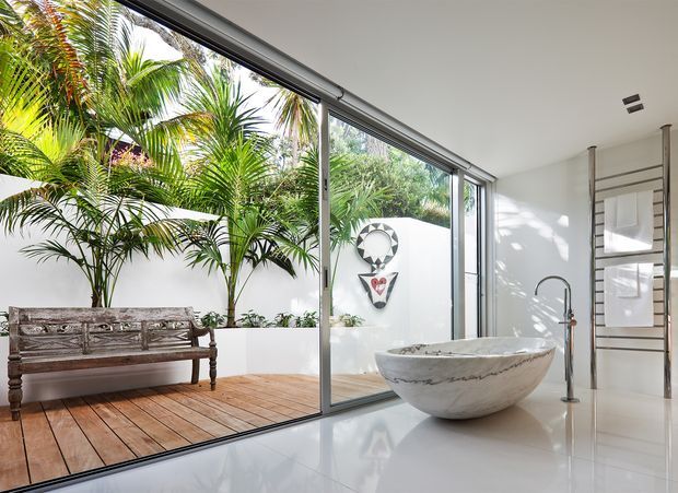 Bright contemporary bathroom with a freestanding white stone bathtub beside full-height sliding glass doors. The glazing opens visually onto a timber deck and lush tropical planting enclosed by a white courtyard wall. A weathered timber bench sits outside, while a sculptural wall feature and ladder-style heated towel rail add detail indoors. The space is highly polished, airy, and strongly connected to an outdoor courtyard.