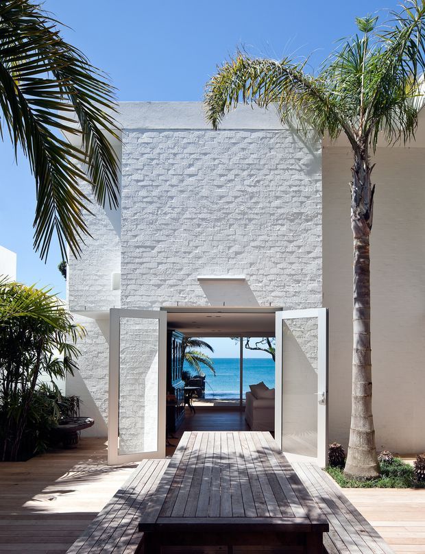 A bright coastal architectural exterior featuring white textured masonry walls, a wide open doorway, timber decking and a long timber table leading toward a blue ocean view. Tall palms and tropical planting frame the entry, while a glimpse of an interior living area is visible beyond the doors.
