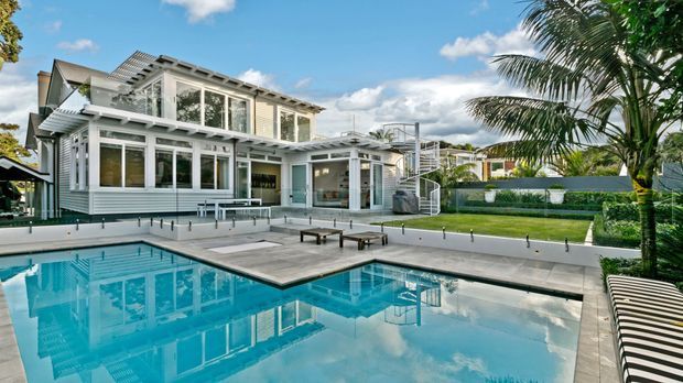 A bright contemporary multi-level residence viewed from beside a large swimming pool. The home features extensive white-framed glazing, horizontal weatherboard-style cladding, projecting roof forms, balconies and outdoor stairs. The pool terrace includes a pale paved deck, sun loungers and manicured hedging, with tropical palm planting around the site.