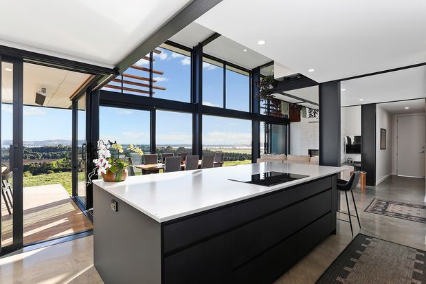 Bright contemporary kitchen with a large white island and black integrated cabinetry, set beside expansive black-framed floor-to-ceiling glazing. The glazing opens toward a timber outdoor deck and landscaped views, while a dining area, pendant-free open-plan space, and hallway are visible beyond. The room has a clean, refined palette of white, black, grey, and warm timber accents.
