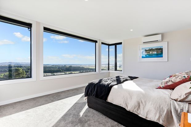 Bright contemporary bedroom with a large bed, soft grey carpet, pale walls and extensive corner glazing overlooking a broad landscape and water view. The room includes a wall-mounted air conditioner, framed artwork, and layered neutral bedding with dark and patterned accents.