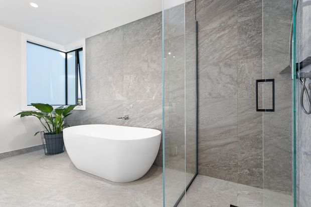 Contemporary bathroom featuring a freestanding white oval bathtub, large-format grey stone-look wall and floor tiles, a frameless glass shower enclosure with dark hardware, and a black-framed window. A potted plant adds a natural accent.