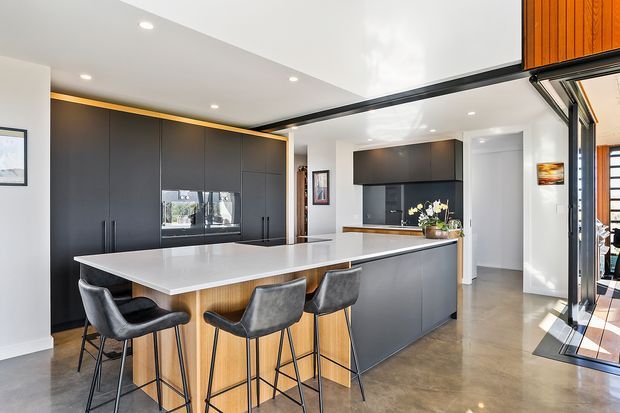 Contemporary open-plan kitchen with a large white island and four dark upholstered bar stools. The kitchen features matte charcoal cabinetry, warm timber accents, integrated appliances, a dark splashback, polished concrete flooring, recessed ceiling lighting, and extensive glazed doors opening to an outdoor area.