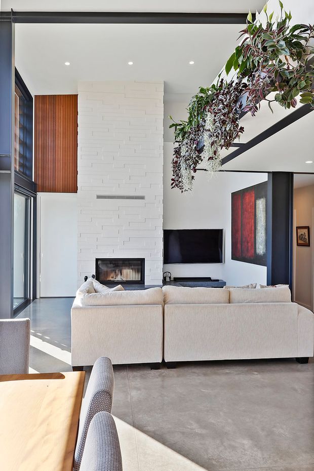 A bright, double-height contemporary living room viewed from behind a large cream sectional sofa. The space features a tall white textured brick fireplace wall with a built-in fireplace, black-framed floor-to-ceiling glazing, dark structural beams, a wall-mounted television, a timber vertical-slat feature wall, polished concrete flooring, and hanging greenery. A dining table and chairs are visible in the foreground.