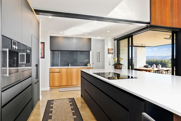 A bright, contemporary kitchen with a large black island topped in white, integrated dark cabinetry and appliances along the left wall, and a timber-fronted rear cabinet run with a dark splashback. Full-width sliding glass doors open to an outdoor dining area and a distant mountainous landscape. Recessed ceiling lights and timber ceiling detailing add warmth and contrast.
