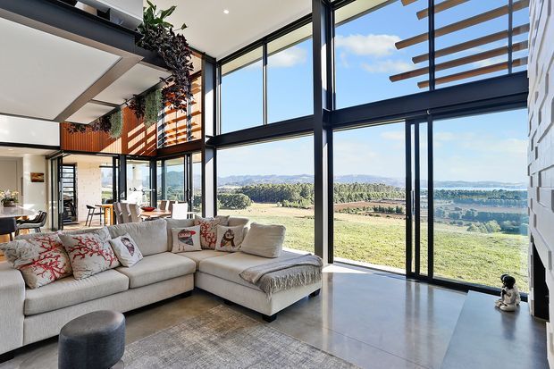 A bright, spacious contemporary living room with a large sectional sofa, expansive black-framed floor-to-ceiling glazing, polished concrete flooring, and a dramatic double-height window wall. The room opens to panoramic rural and coastal views, with timber architectural screens and hanging greenery adding warmth.