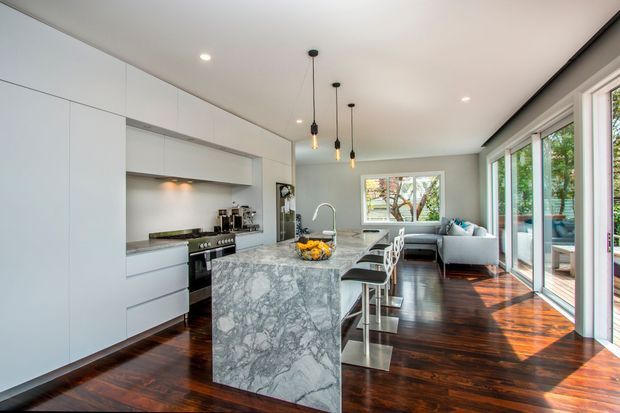 Bright open-plan kitchen and living area with sleek white full-height cabinetry, a large veined stone island with integrated seating, stainless steel appliances, pendant lighting, and rich dark timber flooring. Floor-to-ceiling glazed doors open toward an outdoor deck, while a compact living area sits beyond the kitchen. The space has a clean contemporary feel with strong natural light and warm timber contrast.
