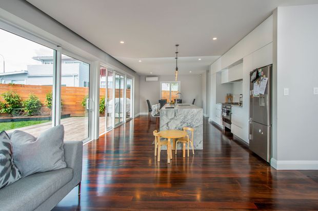 Bright open-plan kitchen and living area with polished dark timber flooring, light grey walls, extensive sliding glass doors, white built-in cabinetry, stainless-steel appliances, and a marble-look kitchen island. A compact timber children's table and chairs sit in the foreground, with a dining area beyond and a timber-fenced outdoor courtyard visible through the glazing.