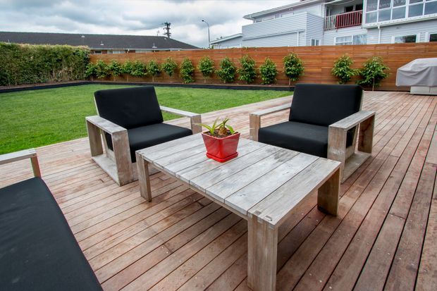 A spacious outdoor entertaining area with a weathered timber deck, two black-cushioned timber armchairs, a matching coffee table, and a partially visible lounge. The deck opens onto a manicured lawn enclosed by a timber privacy fence with planted greenery. A light-coloured, multi-level residential building is visible beyond the fence under an overcast sky.