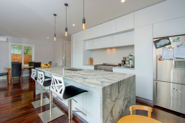 Bright contemporary kitchen with a large waterfall-edge marble island, integrated white cabinetry, stainless-steel appliances, pendant lighting, and bar seating. Dark timber flooring extends toward a dining area and window at the rear.