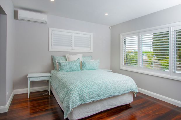 Bright, compact bedroom with a bed dressed in a pale aqua geometric patterned duvet and matching pillows, set against soft grey walls. White plantation-shutter windows provide abundant daylight, while dark polished timber flooring and simple white bedside furniture complete the room. A wall-mounted air-conditioning unit is visible above the bed.