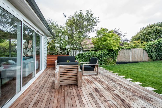 A landscaped residential backyard with a spacious timber deck adjoining a glazed, weatherboard-clad extension. The deck is furnished with a small outdoor seating arrangement, while hedges, mature trees, lawn, and a timber boundary fence surround the outdoor area.