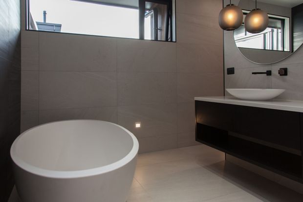 Contemporary bathroom featuring a freestanding white bathtub, large-format grey wall and floor tiles, a floating dark vanity with a white vessel basin, black wall-mounted fixtures, a curved mirror, and two warm globe pendant lights. High windows provide natural light.