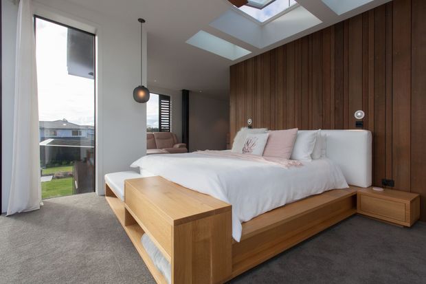 Contemporary bedroom with a low-profile built-in timber bed and integrated bedside cabinetry against a full-height dark timber feature wall. The room has grey carpet, white walls and bedding, soft blush accents, pendant lighting, tall glazing, and multiple skylights that provide abundant natural light.