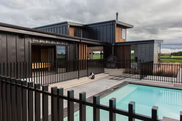 A contemporary dark-clad two-storey residence viewed from the pool terrace. The house features bold vertical metal cladding, timber screening accents, extensive glazing, and a low-pitched roofline. A turquoise swimming pool occupies the foreground, enclosed by a black vertical-bar fence, with paved outdoor areas surrounding it.
