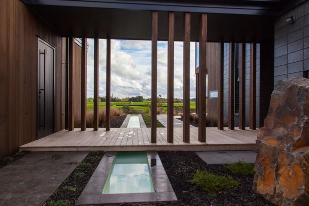 A dramatic modern entryway with dark timber wall cladding, a wide timber deck, and a screen of vertical timber posts framing views across a rural landscape. Large stonework forms a feature element at the right, while a narrow reflective water channel leads toward the entrance.
