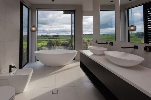 Contemporary bathroom with a freestanding white oval bathtub, bidet, double vessel basins on a dark floating vanity, large mirrors, pendant lights, and expansive windows overlooking green rural fields.