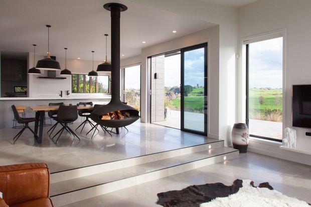 Bright open-plan contemporary living and dining space with a kitchen visible at the rear. The room features polished light flooring, a raised dining platform, expansive black-framed glazing, a suspended black fireplace, pendant lighting, a timber dining table with dark chairs, and views of open green landscape outside.