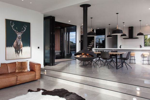 Open-plan contemporary living and kitchen area with a tan leather sofa, sculptural suspended wood-burning fireplace, large dining island with pendant lights, dark-framed glazing, and a minimal white kitchen. The living area is slightly lowered from the kitchen and dining zone, with polished grey flooring and a black-and-white hide rug.