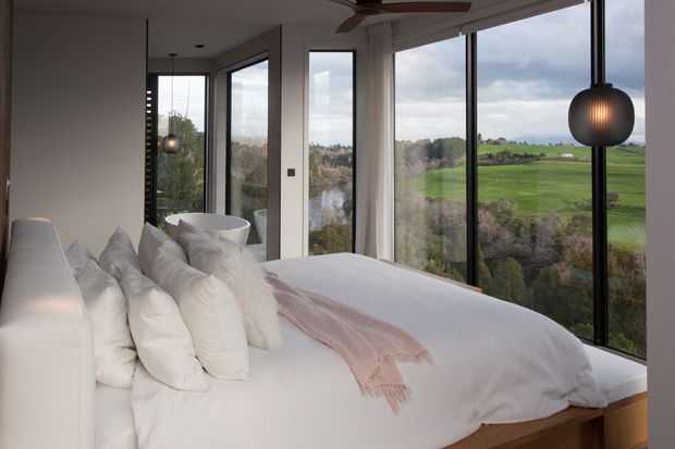 A bright contemporary bedroom with a large white upholstered bed layered with white pillows and a soft blush throw. Floor-to-ceiling black-framed glazing wraps the room, overlooking a green rural landscape and waterway. A spherical pendant light, ceiling fan, and minimalist white furnishings complete the calm interior.