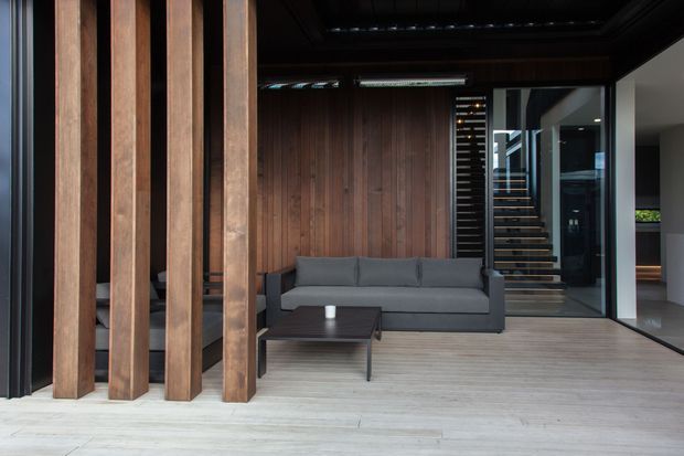 A dark, refined contemporary living room with a grey sofa and low black coffee table set against a vertical timber feature wall. Dramatic timber posts frame the space, while full-height glazing reveals a stairway and adjacent interior areas. Pale timber flooring contrasts with the dark ceiling, walls, and joinery.