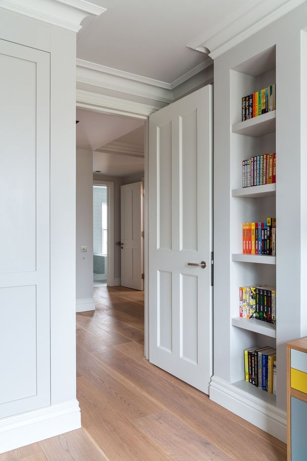 A bright, refined residential hallway with warm timber flooring, white paneled doors, built-in open book shelving, and extensive decorative cornices and skirting boards. The hallway opens toward additional rooms and a bathroom or wet area in the distance.