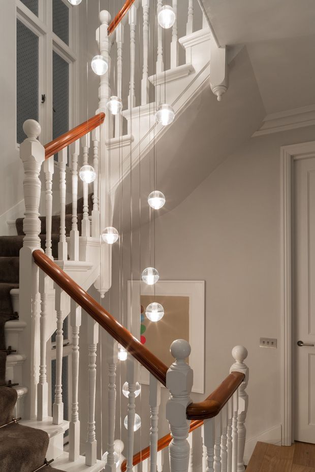 A refined interior stairway with traditional white-painted turned balusters, newel posts, decorative mouldings and warm polished timber handrails. A dramatic cluster of suspended spherical pendant lights follows the stair void, with grey carpeted steps, neutral painted walls, framed artwork, a white panelled door and timber flooring.