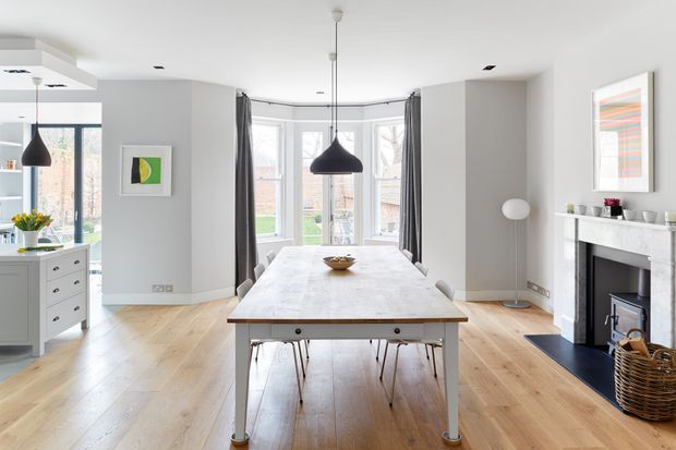 Bright, open-plan dining area with a large pale timber dining table, pendant lighting, light timber flooring, white cabinetry, and grey walls. The room connects visually to a kitchen at left and a living area with fireplace at right, with large bay-style glazing and dark curtains at the rear.