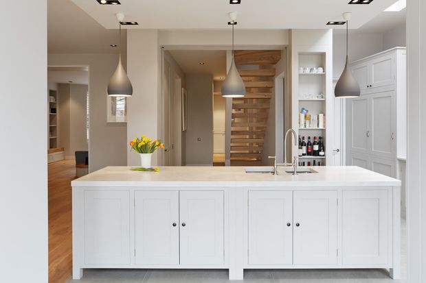 Bright, refined contemporary kitchen centred on a large white island with paneled cabinetry, a pale stone benchtop and an undermount sink. Three grey pendant lights hang above the island, while recessed ceiling lights provide additional illumination. Built-in open shelving displays books and bottles, and a timber open-riser staircase is visible beyond the kitchen. Warm timber flooring and neutral walls create a balanced, inviting interior.