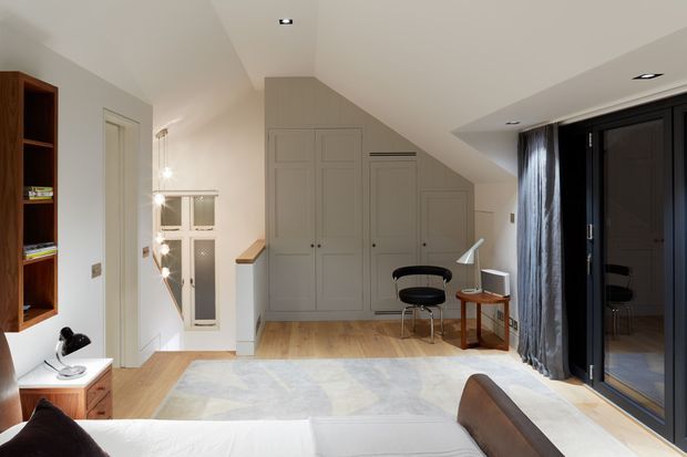 Bright contemporary bedroom with a sloping ceiling and built-in grey cabinetry beneath the eaves. The room includes a partially visible bed, desk and chair, timber flooring, a pale area rug, recessed lighting, and full-height dark-framed glazing with curtains leading to an exterior or balcony area.