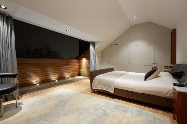 A spacious, refined bedroom with a low-profile upholstered bed, white bedding, expansive dark glazing, warm horizontal timber wall panelling, built-in white wardrobes, and recessed ceiling lighting. A large pale area rug and neutral finishes create a calm, softly lit atmosphere.
