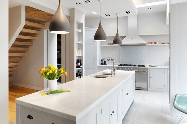 Bright contemporary kitchen with a large white island, integrated sink and white paneled cabinetry. The space includes a stainless-steel range and hood, pale stone surfaces, a textured grey splashback, timber open-riser staircase, and three sculptural pendant lights. A small dining or seating area is visible at the right edge.